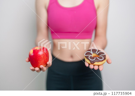 woman hand hold red Apple and donut, female fitness choose between fruit is Healthy and sweet is Unhealthy junk food. Dieting control, Weight loss, Obesity, eating lifestyle and nutrition concept 117735904