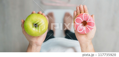 woman on weight scale and hand hold green Apple and donut, choose between fruit is Healthy and sweet is Unhealthy junk food. Dieting control, Weight loss, Obesity, eating lifestyle and nutrition 117735920