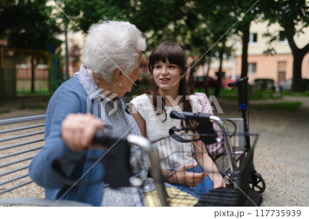 Grandma with walker spending time with granddaughter in city park, sitting on bench at children playground, talking. After school. 117735939