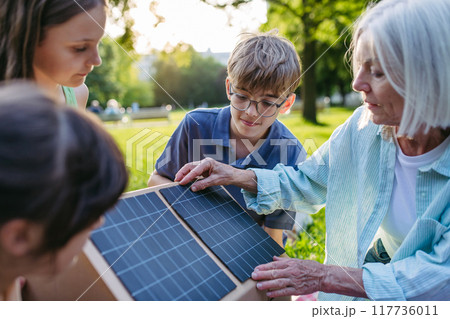 Children learning about renewable energy and solar panels during sustainable education class outdoors, using cardboard model of house wit solar panel on roof. 117736011
