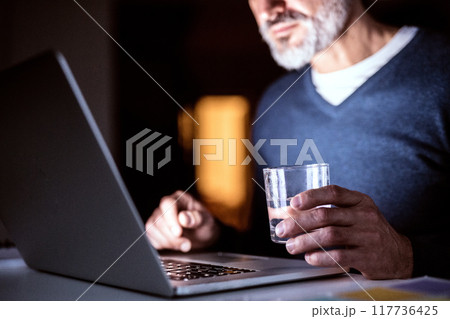 Handsome man drinking while working from home office at evening, using laptop. Screen glow illuminates man's face. Telecommuting and home office. 117736425