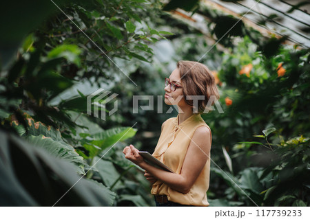 Small greenhouse business. Businesswoman selling flowers and houseplants, holding tablet and smiling. Small greenhouse business. Businesswoman selling flowers and houseplants, holding tablet and smiling. 117739233