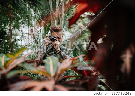 Handsome man with tattoos standing in botanical garden, in the middle of lush green foliage, holding camera and taking photography. 117739248