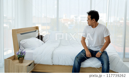 Man sitting on edge of bed looking over his shoulder, while woman sleeps under white duvet in bright bedroom with large windows Man sitting on edge of bed looking over his shoulder, while woman sleeps under white duvet in bright bedroom with large windows 117739262