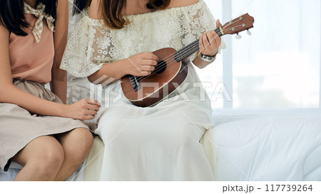 Close-up of two women sitting together, one playing ukulele with focus on her hands and instrument, while other gestures beside her, both dressed casually in bright room 117739264