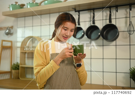 Young woman wearing apron enjoying morning coffee in her kitchen at home Young woman wearing apron enjoying morning coffee in her kitchen at home 117740692