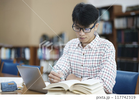 Young Asian male sitting inside a library alone doing research. Man working on a project. Young man doing research for a case. Young Asian male sitting inside a library alone doing research. Man working on a project. Young man doing research for a case. 117742319