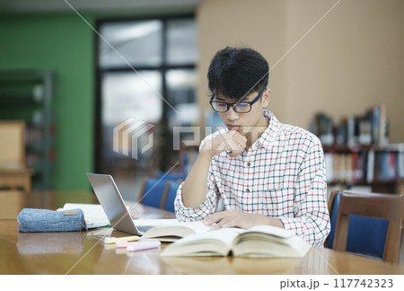 Young Asian male sitting inside a library alone doing research. Man working on a project. Young man doing research for a case. Young Asian male sitting inside a library alone doing research. Man working on a project. Young man doing research for a case. 117742323