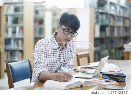 Young Asian male sitting inside a library alone doing research. Man working on a project. Young man doing research for a case. Young Asian male sitting inside a library alone doing research. Man working on a project. Young man doing research for a case. 117742327