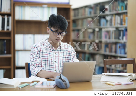 Young Asian male sitting inside a library alone doing research. Man working on a project. Young man doing research for a case. 117742331