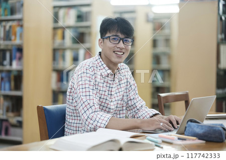 Young Asian male sitting inside a library alone doing research. Man working on a project. Young man doing research for a case. 117742333