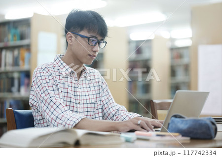 Young Asian male sitting inside a library alone doing research. Man working on a project. Young man doing research for a case. 117742334