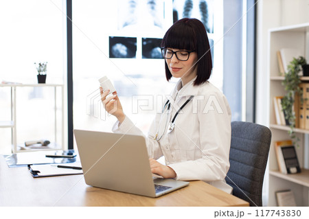 Female doctor working on laptop and holding medicine bottle 117743830