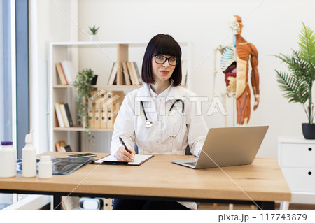 Female doctor taking notes while working on laptop in office 117743879