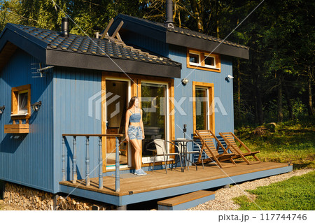 Woman leaning against the doorway of cozy tiny house surrounded by trees in a sunny forest clearing Woman leaning against the doorway of cozy tiny house surrounded by trees in a sunny forest clearing 117744746