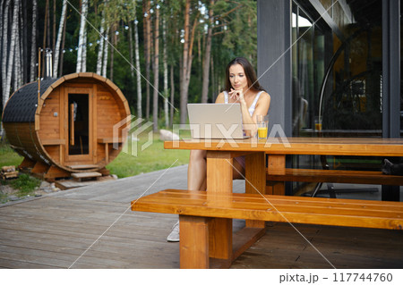 Woman watching movie on a laptop while sitting on a wooden bench on the terrace of a country house Woman watching movie on a laptop while sitting on a wooden bench on the terrace of a country house 117744760