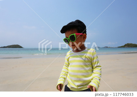 A boy about two years old wearing a long-sleeved shirt and wear sunglasses, walking on the beach smiling happily. 117746033