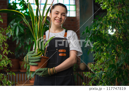 Running of own business. Young woman florist holding plant in pot wearing apron in botanical store. Happy small business owner working at flower shop smiling surrounded by plants. Small business 117747819