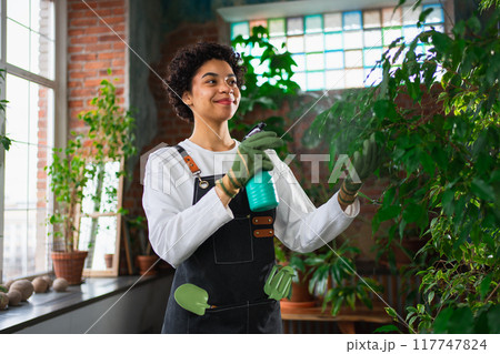 Running of own business. African woman florist with spray bottle watering fertilizing plants in botanical store, Small business owner working at flower shop smiling surrounded by plants Small business 117747824