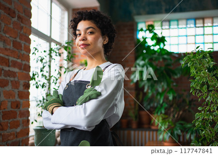 Running of own business. African woman florist wearing apron in botanical store with green plants. Happy small business owner working at flower shop smiling surrounded by plants Small business 117747841