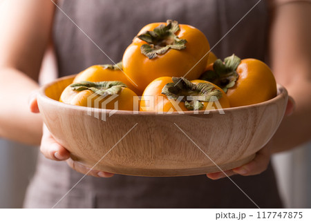 Ripe persimmon fruit in wooden bowl holding by womand hand Ripe persimmon fruit in wooden bowl holding by womand hand 117747875