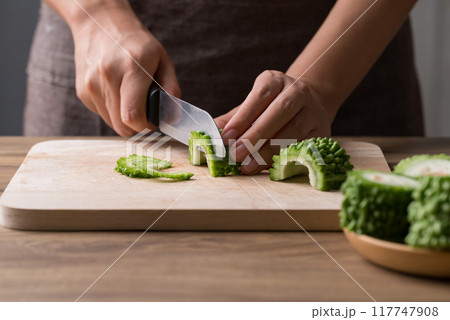 Hand holding kitchen knife and slice bitter gourd on cutting wooden board, Homemade cooking 117747908