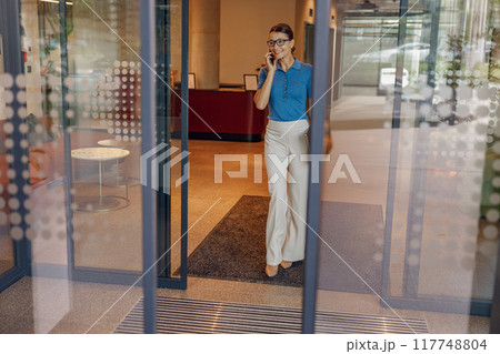A professional businesswoman gracefully leaving her office while engaged with her smartphone 117748804