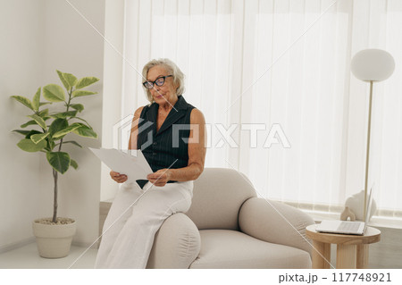 An Elderly Woman Engaged in Reading a Book in an Airy, Bright, and Modern Living Room Space An Elderly Woman Engaged in Reading a Book in an Airy, Bright, and Modern Living Room Space 117748921