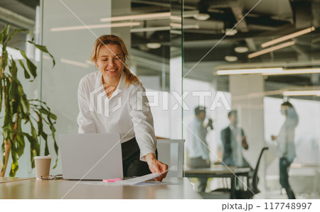 A Smiling Businesswoman is Engaged in Working on Her Laptop Inside a Modern Office Environment A Smiling Businesswoman is Engaged in Working on Her Laptop Inside a Modern Office Environment 117748997