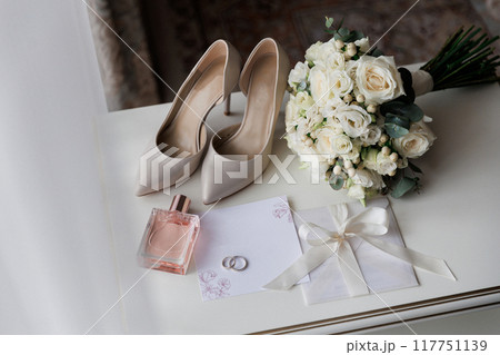 Bride's wedding essentials: heels, bouquet, rings, perfume, and invitation on a white table Bride's wedding essentials: heels, bouquet, rings, perfume, and invitation on a white table 117751139