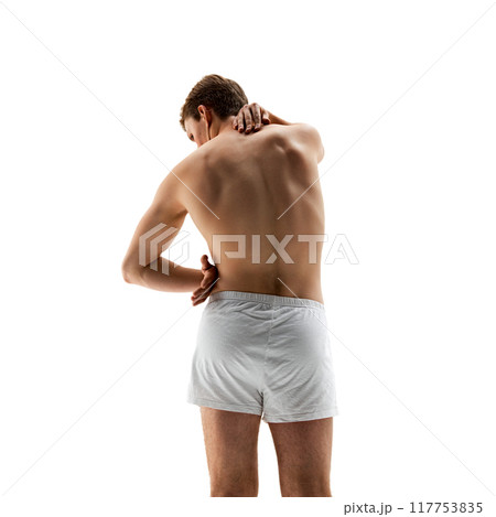 Rear view of man stands in white boxer shorts, with his hand massaging his neck and his right hand holding his lower back, indicating discomfort or pain. 117753835