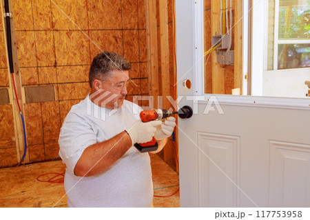 Professional locksmith installing lock on entrance door of house using screwdriver 117753958