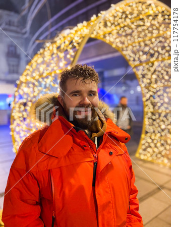 Handsome millennial man model with a hairstyle in winter red jacket walks in night city with festive lights. Christmas and winter holidays season Handsome millennial man model with a hairstyle in winter red jacket walks in night city with festive lights. Christmas and winter holidays season 117754178