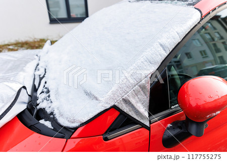 Close-up detail red car with snow-covered windshield protected by reflective cover mat, parked outside residential building during winter. Windscreen window ice frost protection 117755275
