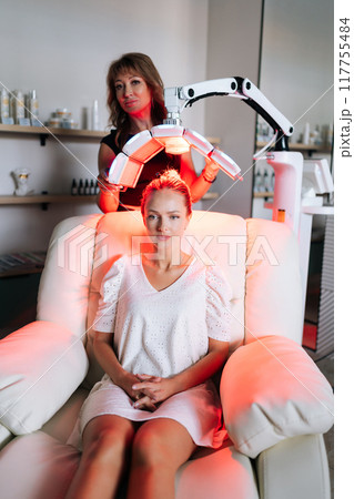 Vertical portrait of beautiful blonde female having LED light hair photodynamic therapy treatment at modern medical clinic. Cosmetician specialist adjusting innovation device for PDT therapy procedure 117755484