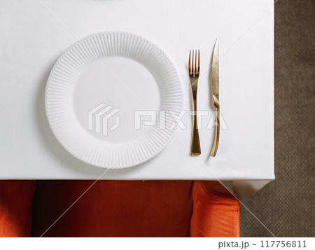 Top view of table in restaurant or dinning room with white round empty plate and gold cutlery. White tablecloth, red chair and table setting as background for your text or design. Copy space 117756811