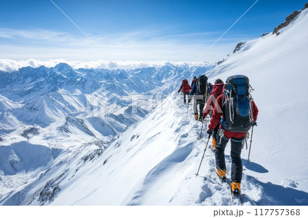 A line of climbers on a snow-covered slope with an expansive view of a dramatic mountain range stretching to the horizon under a clear blue sky 117757368