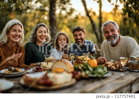 A joyful multi-generational family enjoying an outdoor dinner with a table full of food, laughing and bonding in a natural setting during autumn 117757401