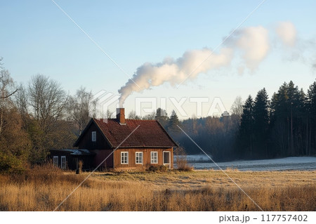 A rustic wooden house with smoking chimney stands in a snowy field, surrounded by bare trees and forest, capturing the essence of rural winter life 117757402