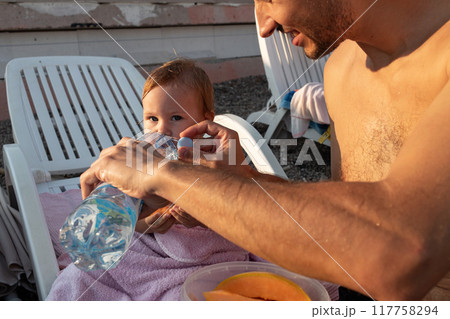 A father gives water from a plastic bottle to his two-year-old daughter after swimming in the sea. A father gives water from a plastic bottle to his two-year-old daughter after swimming in the sea. 117758294