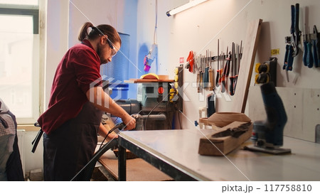 Carpenter collaborating with apprentice, using orbital sander with fine sandpaper to achieve refined finish. Manufacturer and BIPOC colleague using angle grinder on wood block, camera B Carpenter collaborating with apprentice, using orbital sander with fine sandpaper to achieve refined finish. Manufacturer and BIPOC colleague using angle grinder on wood block, camera B 117758810