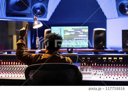 Sound engineer counting down to record singer in sound booth, doing a sign to let the artist know when to start the performance. Audio technician operating control room panel board desk. Sound engineer counting down to record singer in sound booth, doing a sign to let the artist know when to start the performance. Audio technician operating control room panel board desk. 117758816