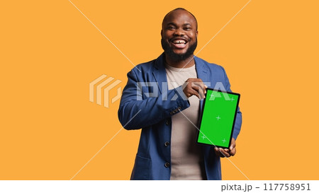 Amused african american man laughing while presenting tablet with green screen display, studio background. Joyous BIPOC person has fun during promotion for blank copy space mockup device, camera B 117758951