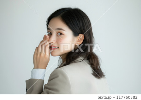 ひそひそ話。耳打ちのポーズをとる20代のアジア人女性。白い背景。ベージュ色のスーツ。 ひそひそ話。耳打ちのポーズをとる20代のアジア人女性。白い背景。ベージュ色のスーツ。 117760582