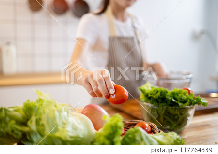 A close-up image of a woman grabbing a tomato while preparing vegetables at the kitchen table. A close-up image of a woman grabbing a tomato while preparing vegetables at the kitchen table. 117764339