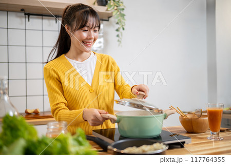 A beautiful Asian woman is cooking in the kitchen, opening a pot lid to check her soup. 117764355