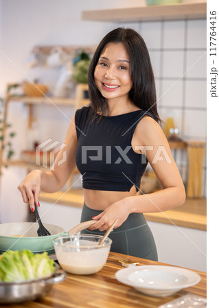 A beautiful, slim Asian woman in workout attire cooking in the kitchen, making pancakes. A beautiful, slim Asian woman in workout attire cooking in the kitchen, making pancakes. 117764416