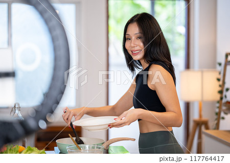 Attractive Asian female food blogger is cooking in front of the camera, recording a video of herself Attractive Asian female food blogger is cooking in front of the camera, recording a video of herself 117764417