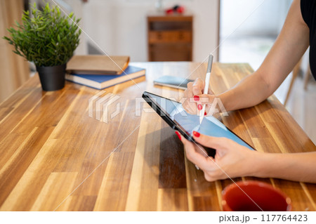 A close-up image of a woman writing on a digital tablet while sitting at the kitchen table. A close-up image of a woman writing on a digital tablet while sitting at the kitchen table. 117764423