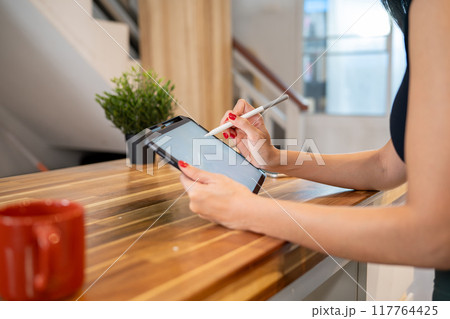 A close-up image of a woman writing on a digital tablet while sitting at the kitchen table. A close-up image of a woman writing on a digital tablet while sitting at the kitchen table. 117764425